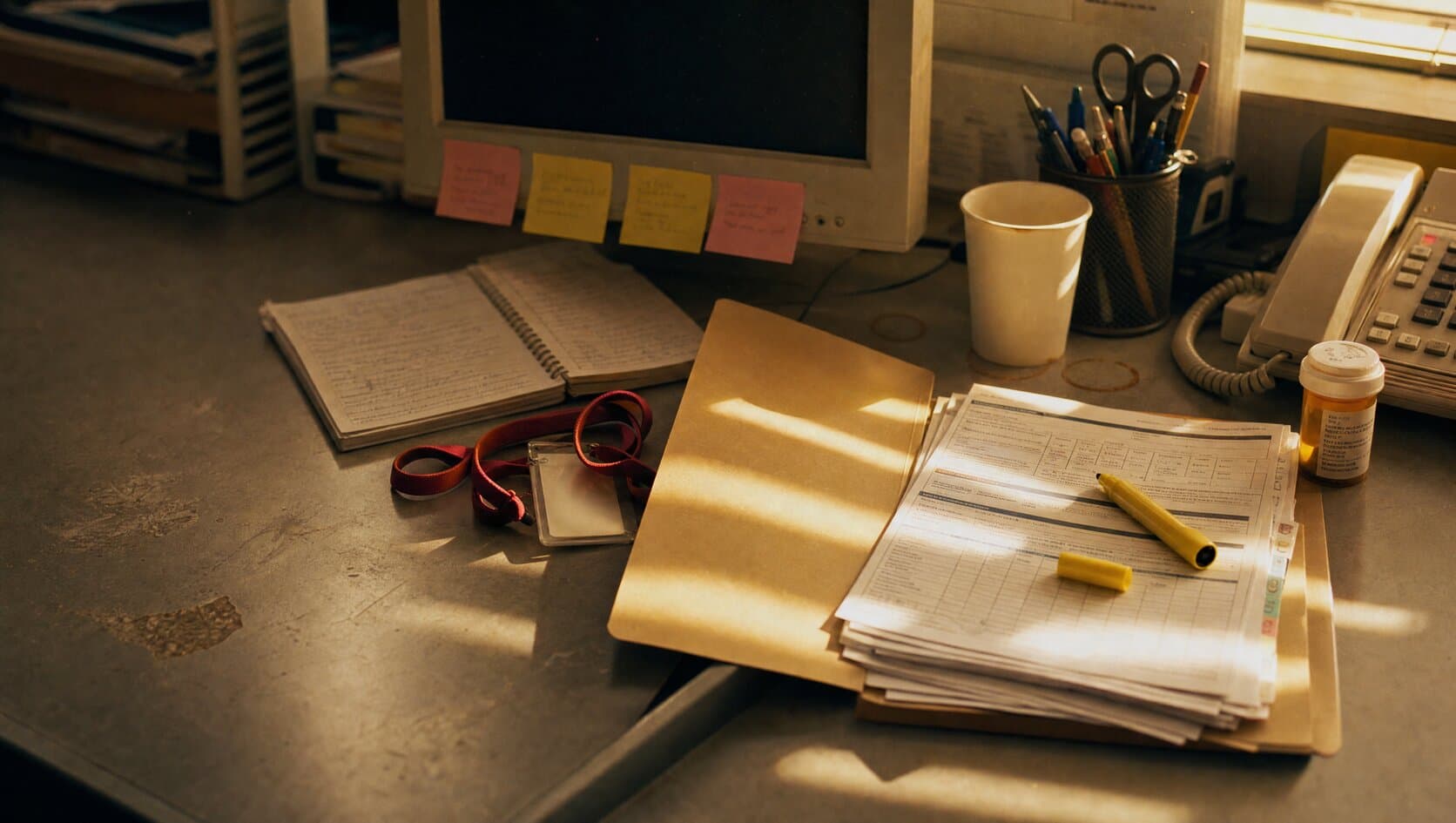 A case manager's desk in late afternoon light, with an open folder of discharge paperwork, a highlighter, a desk phone, and a prescription bottle