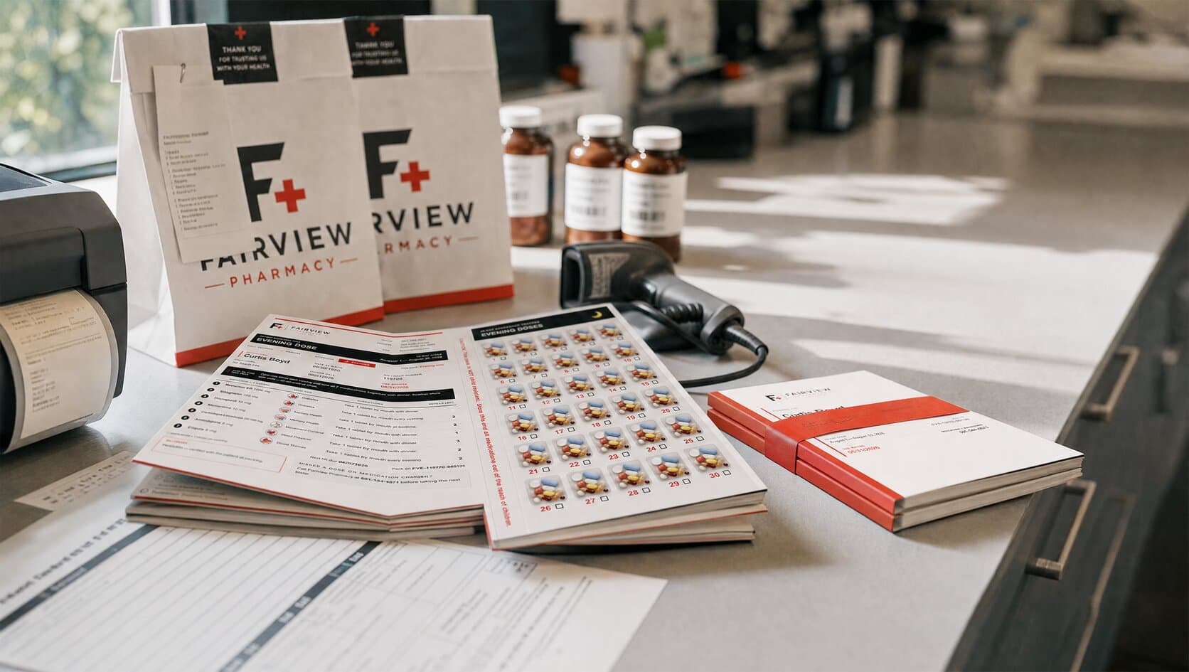The Fairview Pharmacy counter with labeled prescription bags, a strip of sealed multi dose pouches, medication records, and reference cards laid out before a route
