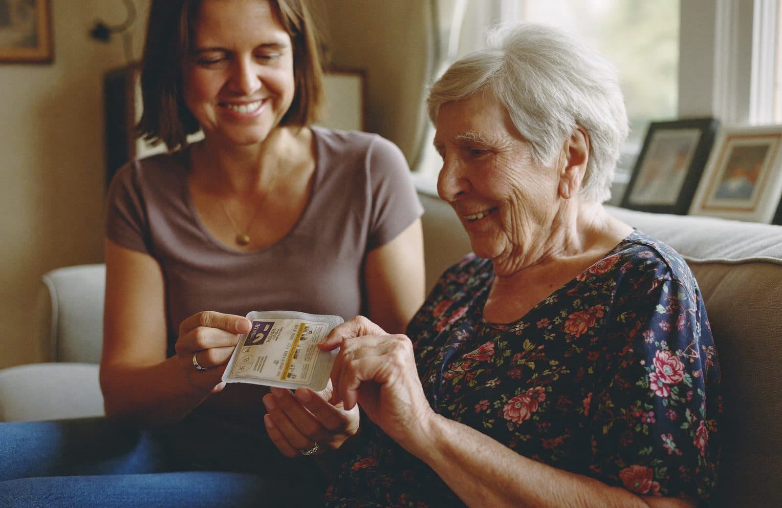 An adult daughter sitting beside her elderly mother on a living room sofa, helping her open a sealed medication pouch in soft window light
