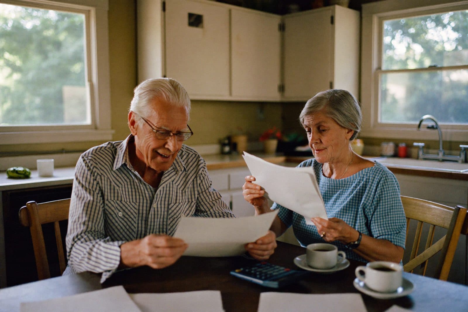 An older couple at their kitchen table in morning light, reviewing plan paperwork together over coffee with a calculator between them