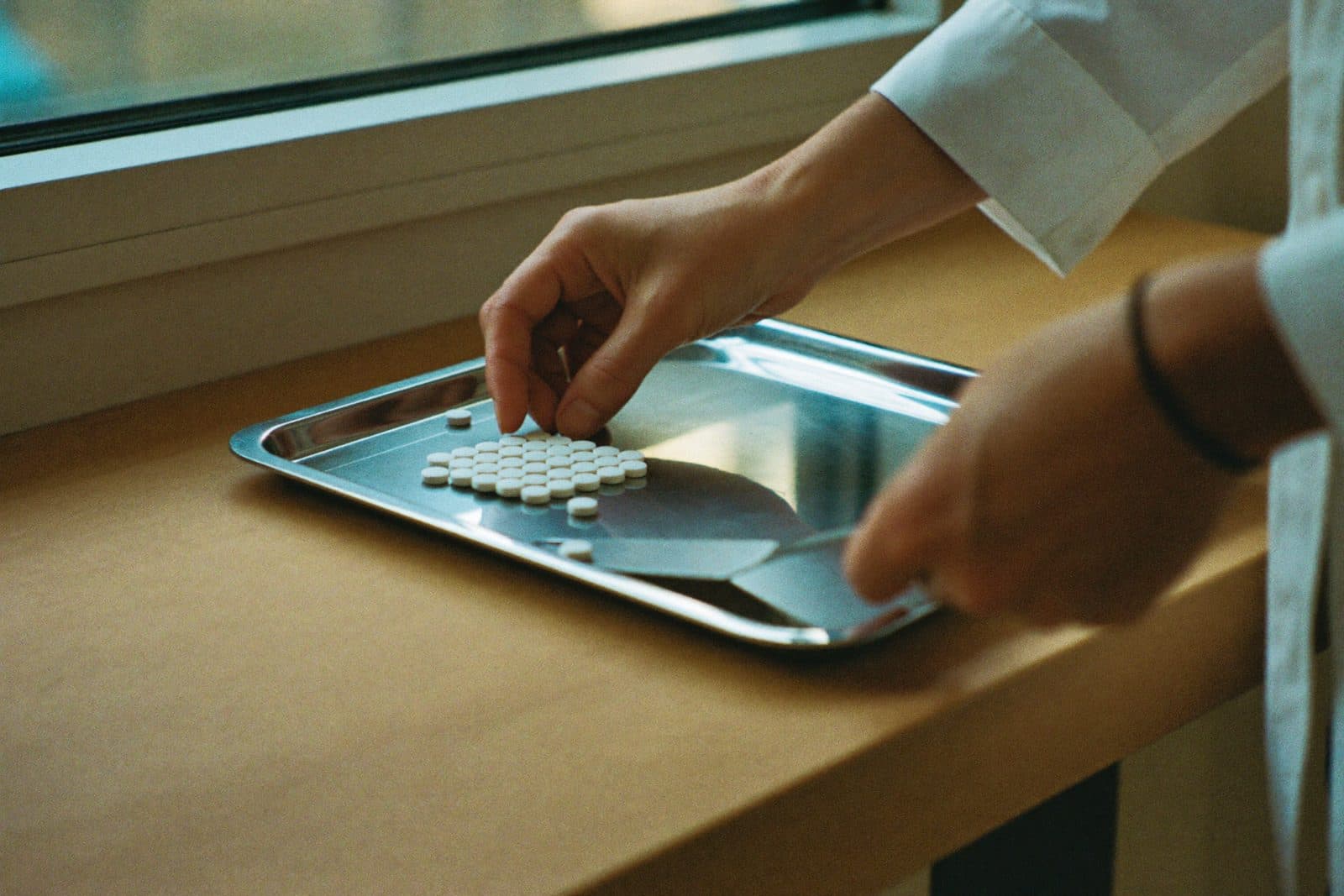A pharmacist's hands arranging white tablets on a stainless steel counting tray at a counter in warm window light