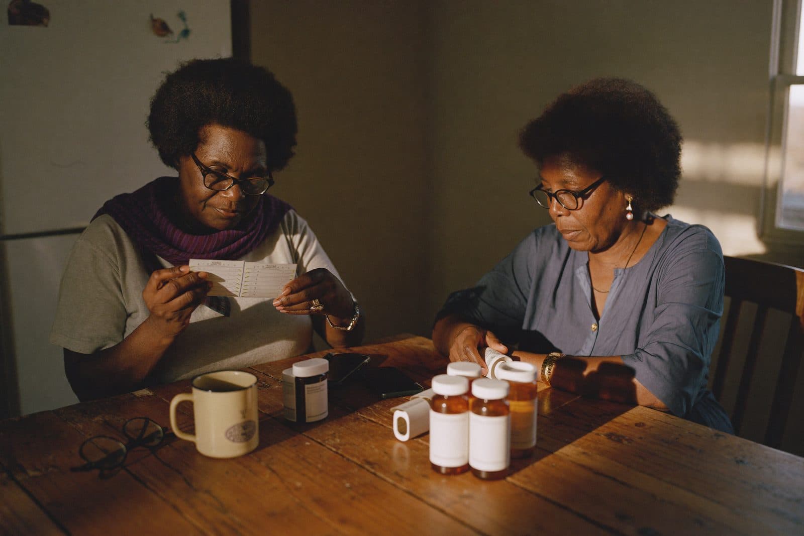 Two women sorting prescription bottles into a weekly pill organizer at a kitchen table in warm afternoon light